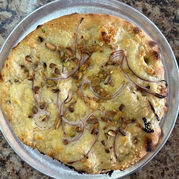 Tony & Alba's focaccia bread with cheese, red onions, and pistachios on a metal pizza pan.