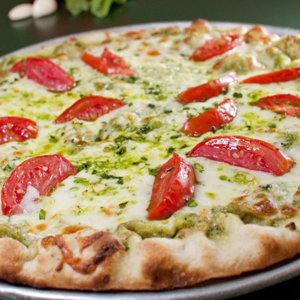 Tony & Alba's pesto pizza with melted mozzarella cheese and sliced red tomatoes on a metal pan.